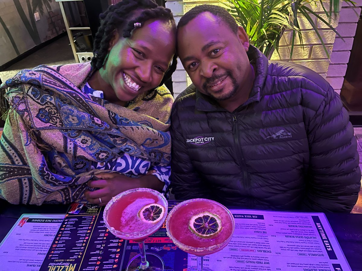 Brian and Manka side by side at a table in the evening, with colourful drinks in front of them.