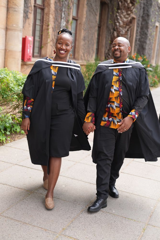 Brian and Manka in their graduation gowns, walking hand in hand and laughing.